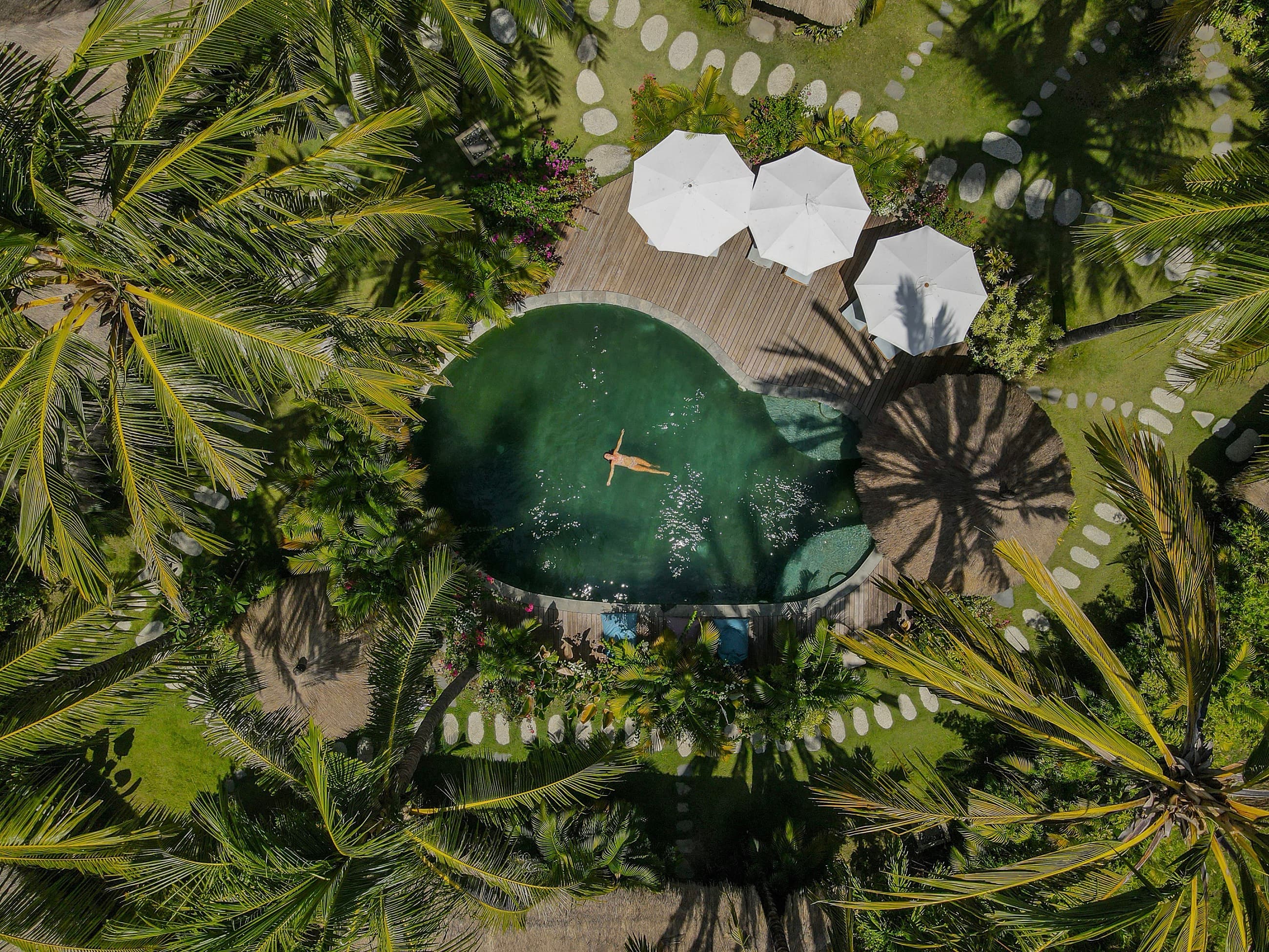 Aerial view of a pool ringed by coconut palms with white parasols on a timber deck, Mana Eco Retreat, Kuta Lombok