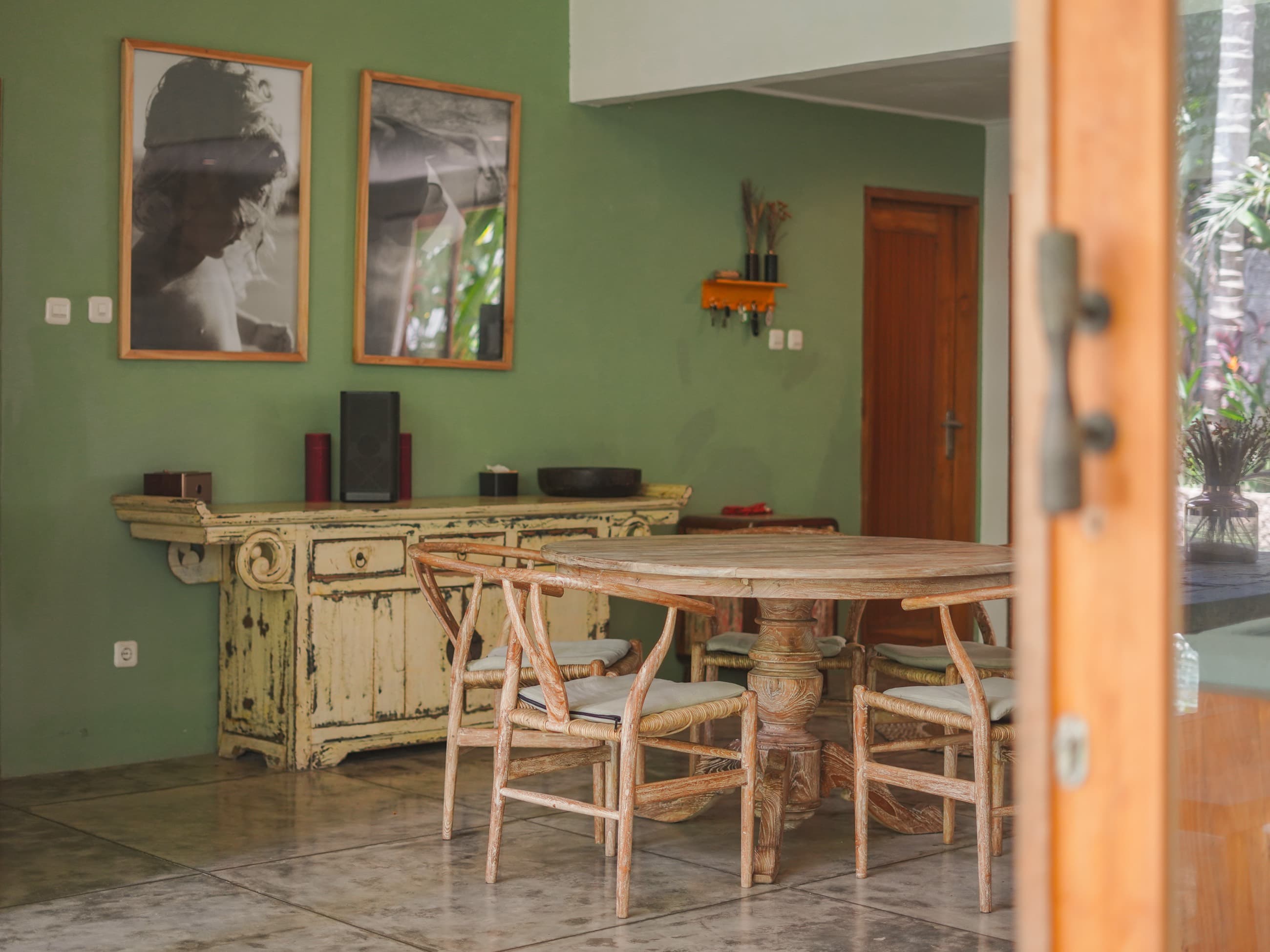 Coin repas de la villa avec table ronde en bois, chaises tressées et cadres sur un mur vert