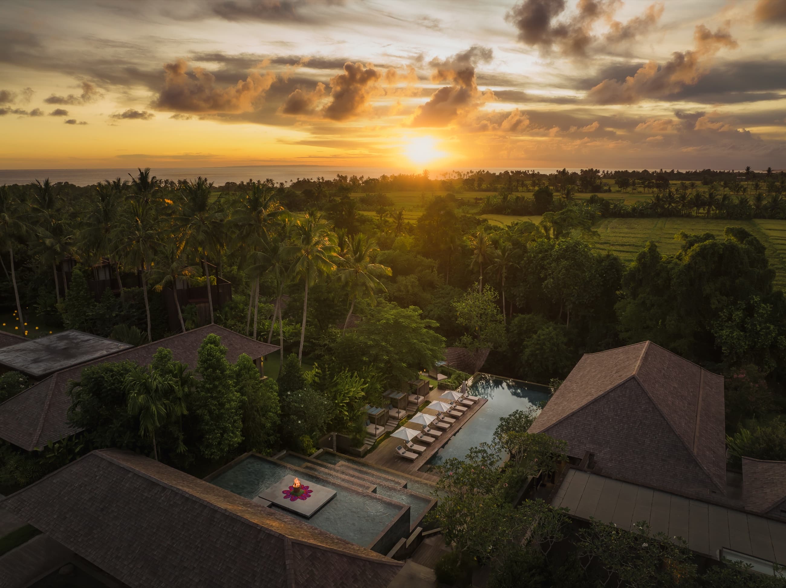 Aerial view at sunset over the pool and villa rooftops, with rice fields and the sea beyond — Nirjhara, Bali