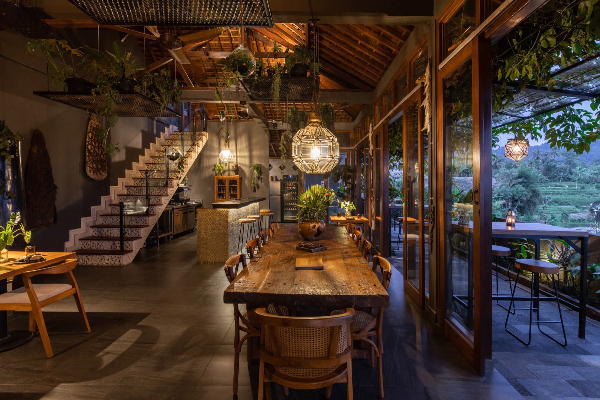 Asri Dining interior — communal wooden table, hanging plants, and warm evening lighting