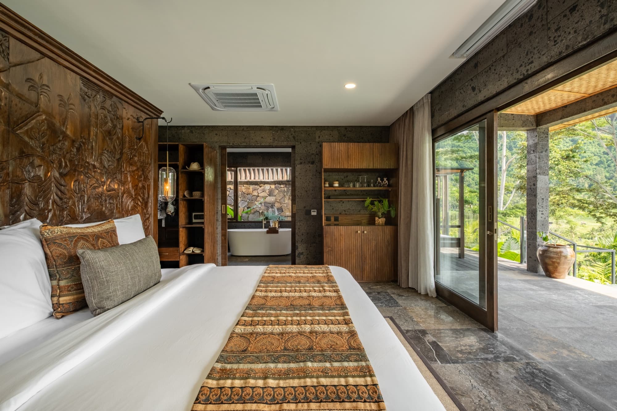 Mulia Wellness Villa bedroom — carved wooden headboard, Indonesian stone, and sliding doors to private pool