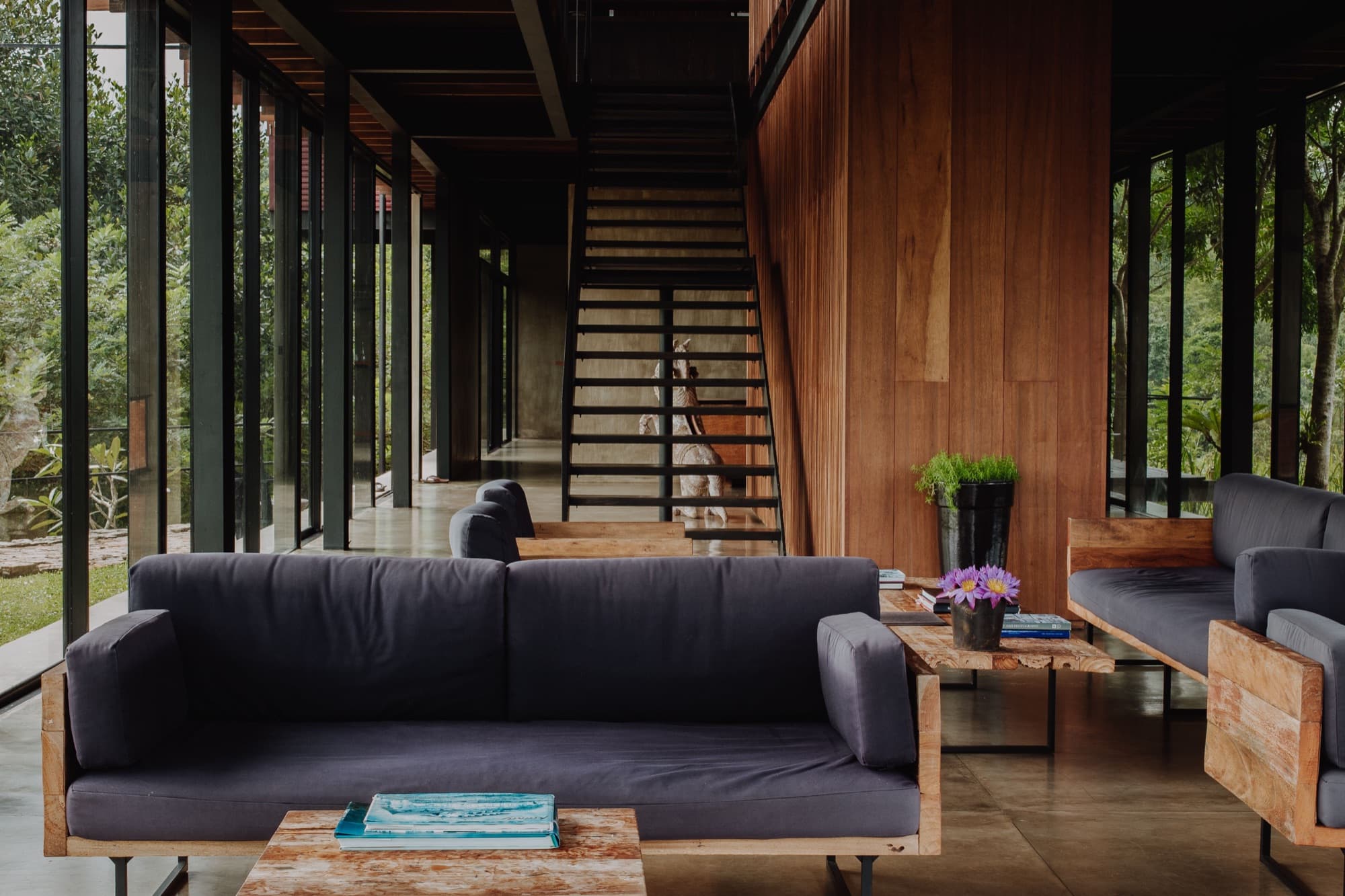 Main pavilion lounge with dark sofas, a floating timber staircase and glass walls onto the forest