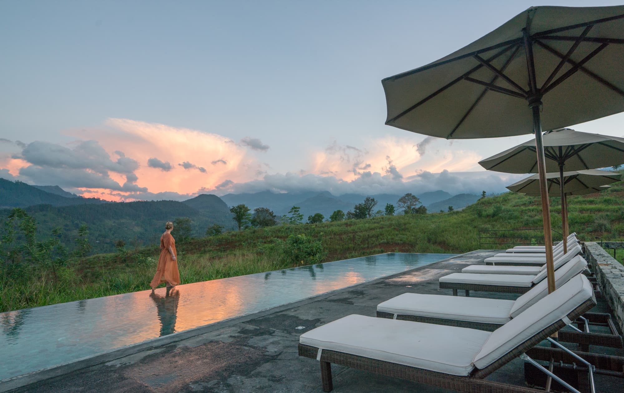 A guest walks the edge of the infinity pool beneath pink sunset clouds over the mountains
