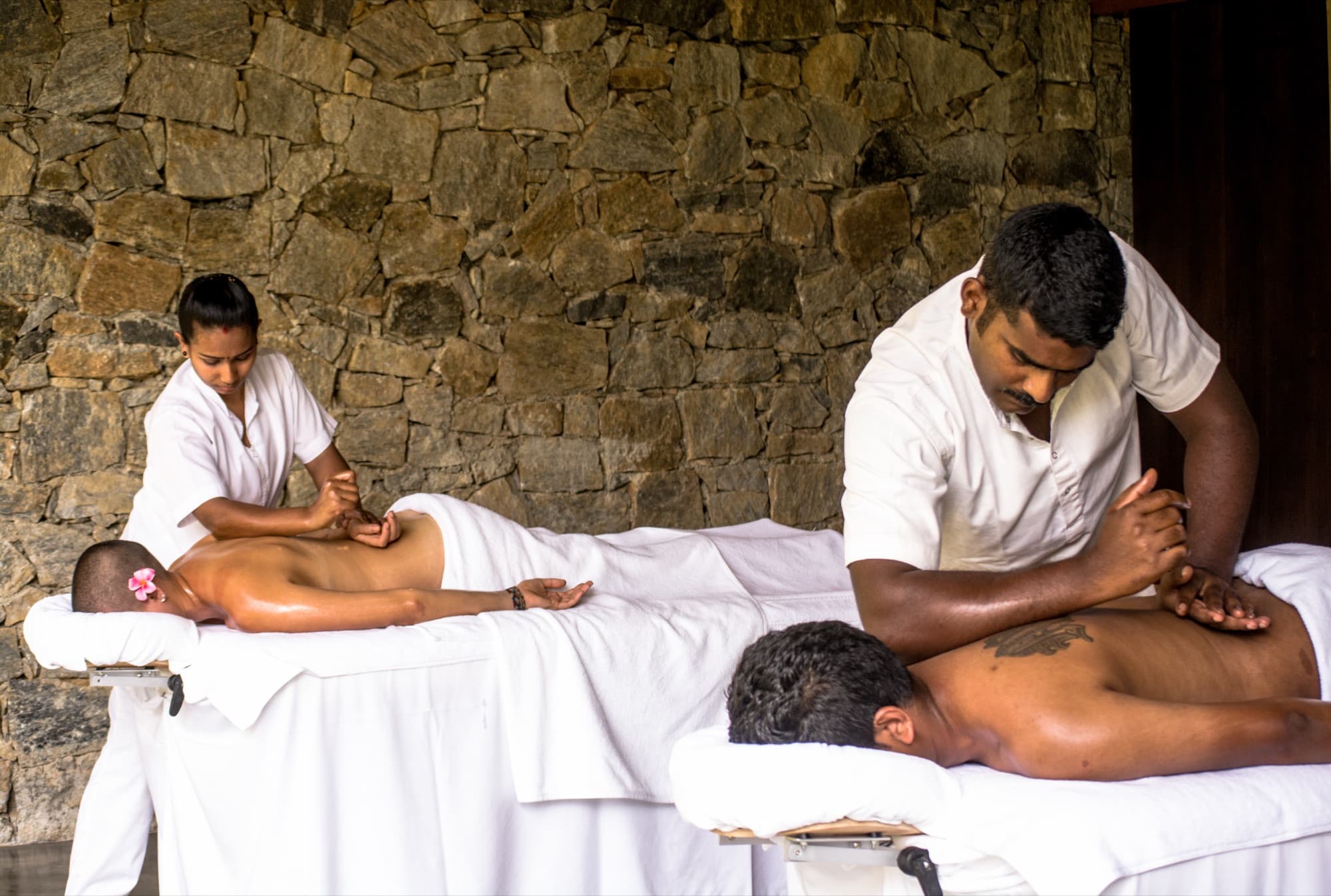 Two guests receiving side-by-side massages from therapists against a stone wall