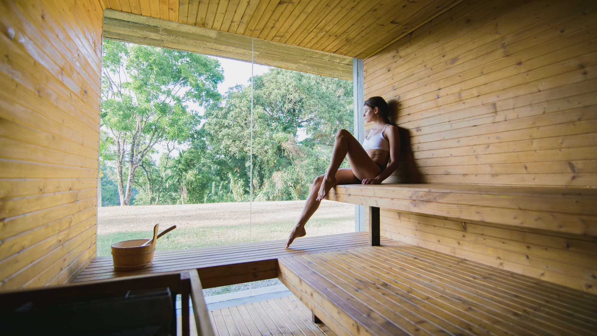 A guest relaxing in the timber sauna beside a large window onto the greenery