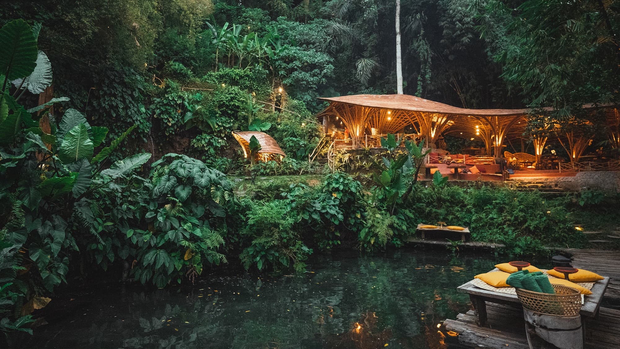 Riverside dining pavilion lit at dusk beside a natural pond