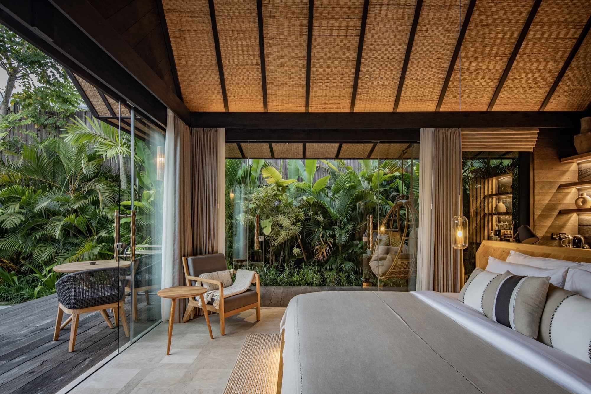Villa bedroom opening through glass walls to the surrounding jungle, with a hanging chair