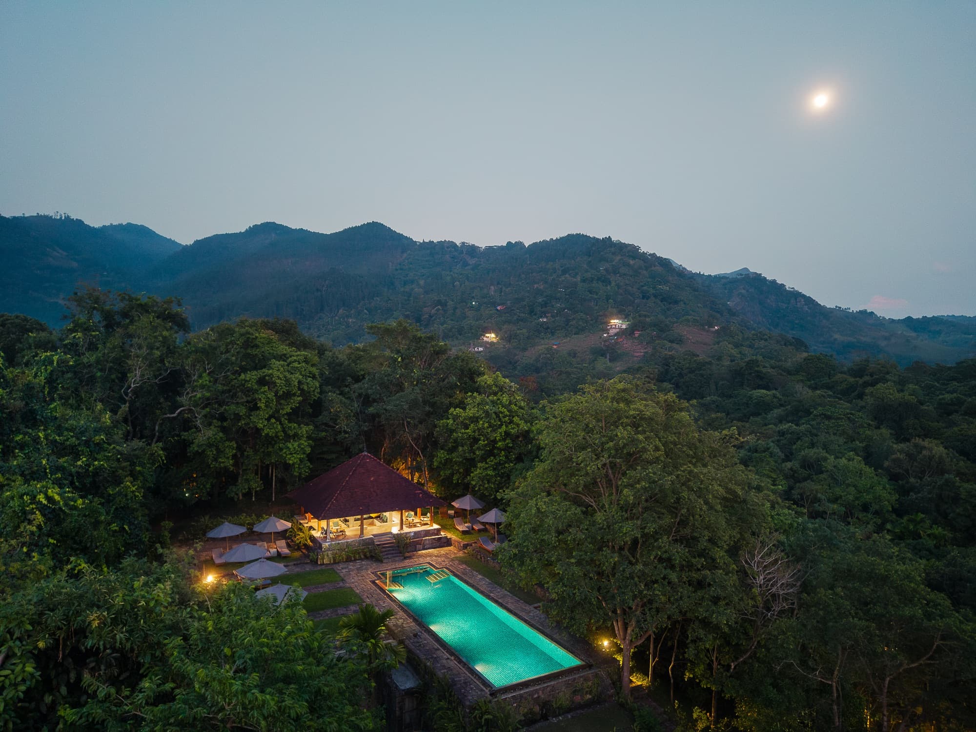 Aerial view of the lit pool at dusk under a rising moon, ringed by rainforest and hills
