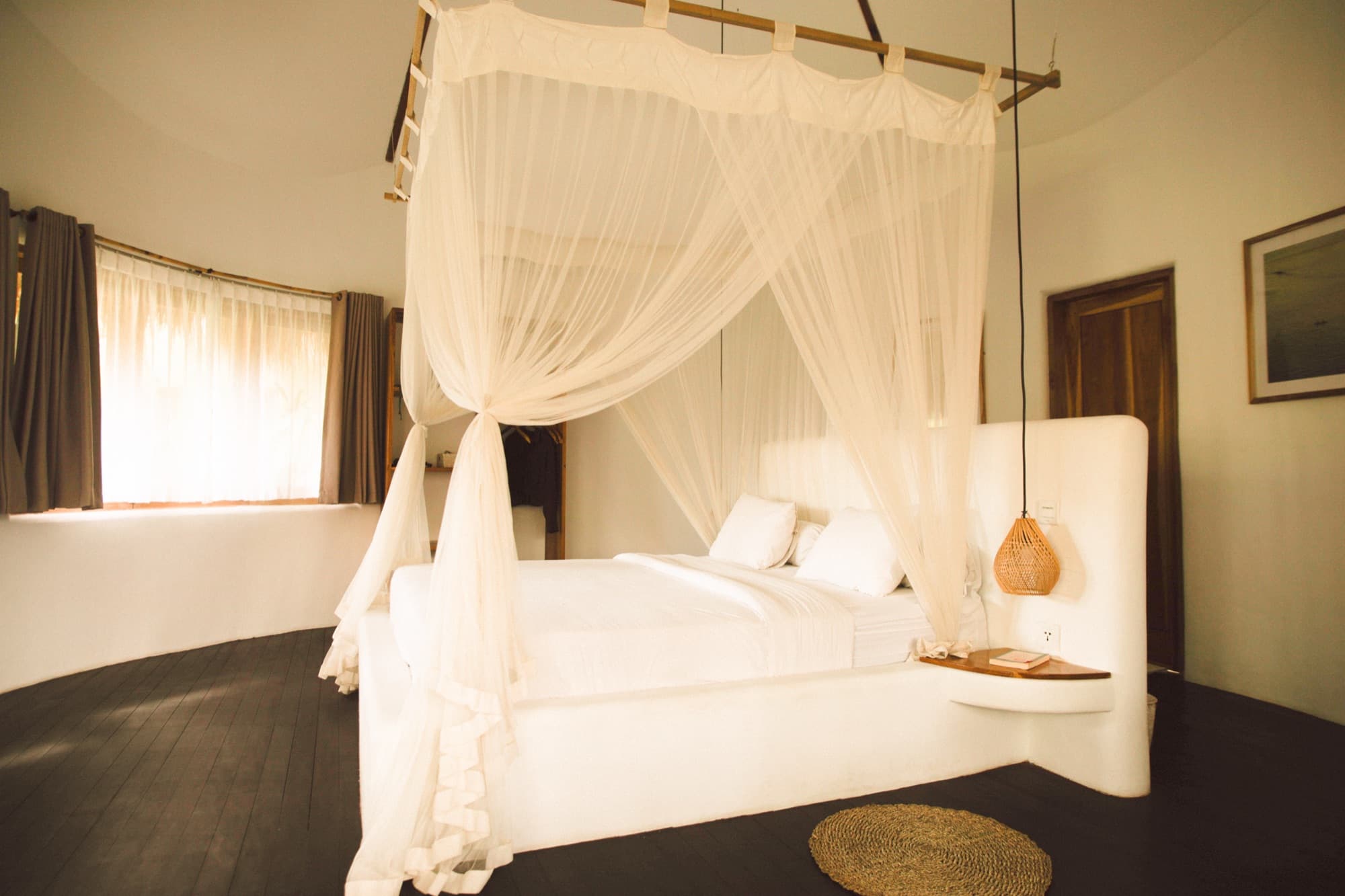 Room with a white canopy bed and mosquito net, dark timber floor and garden window
