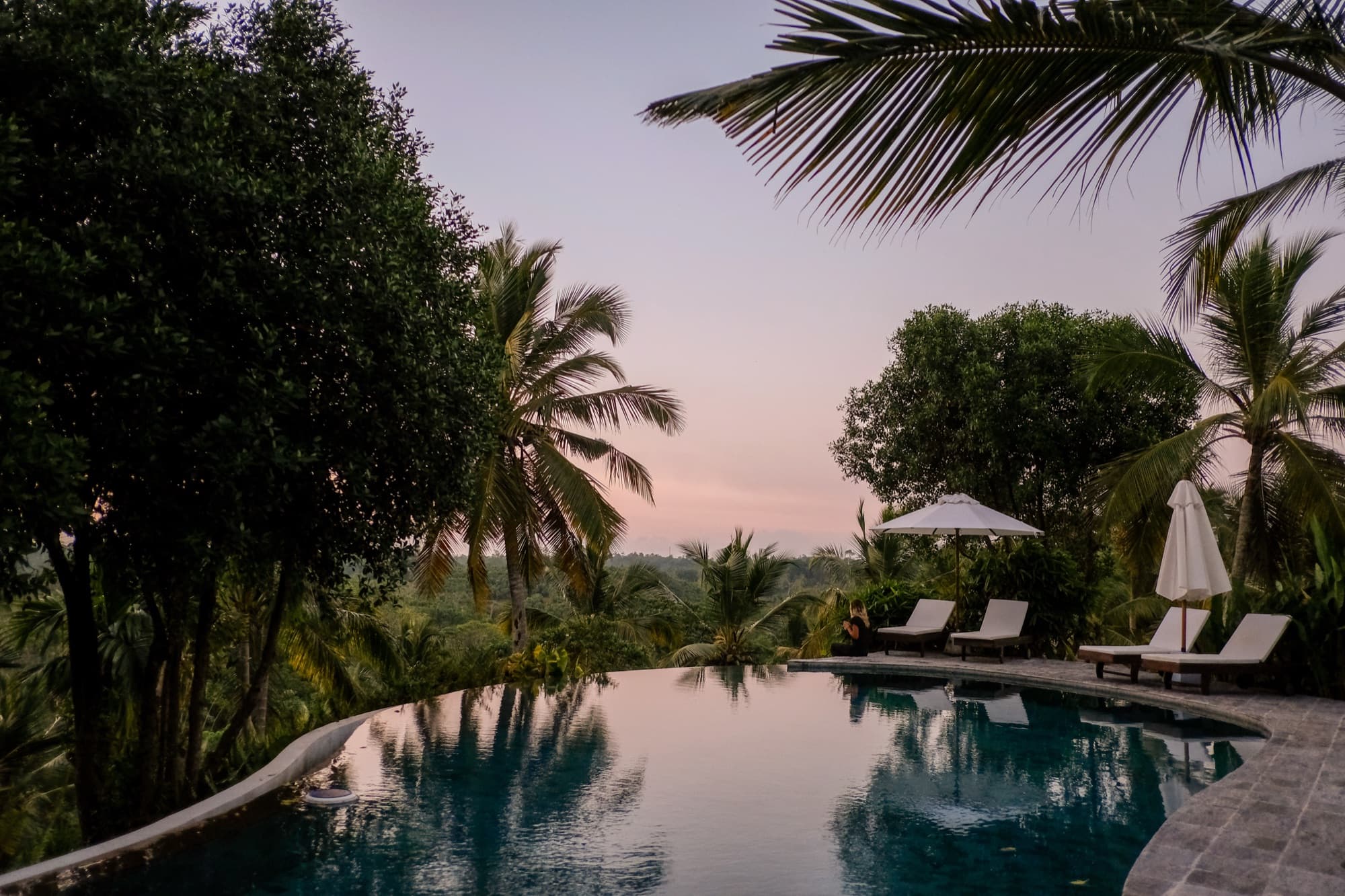 Infinity pool reflecting a pink dusk sky, framed by palms and loungers