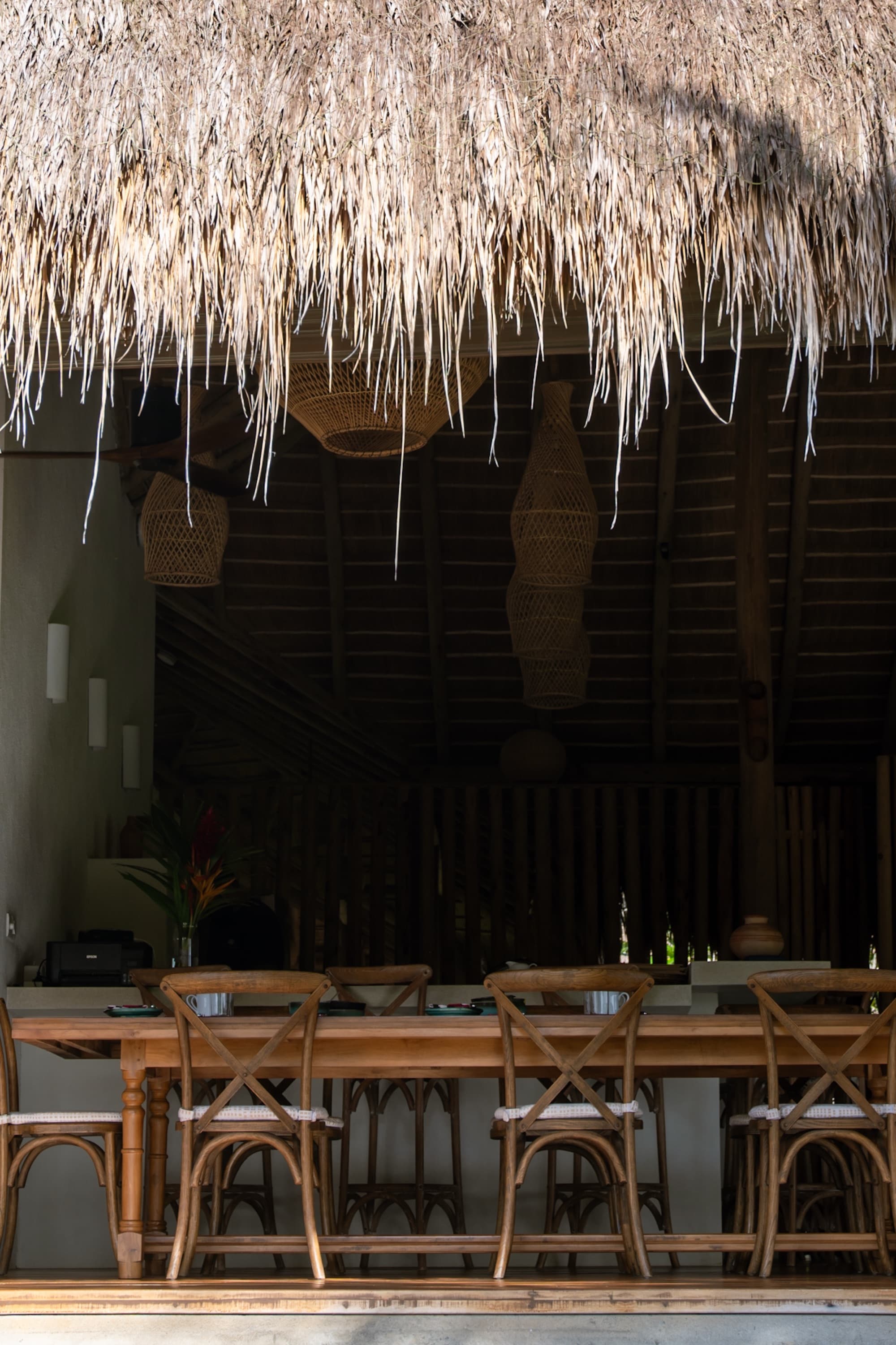 Long communal dining table under a thatched roof with rattan pendant lights