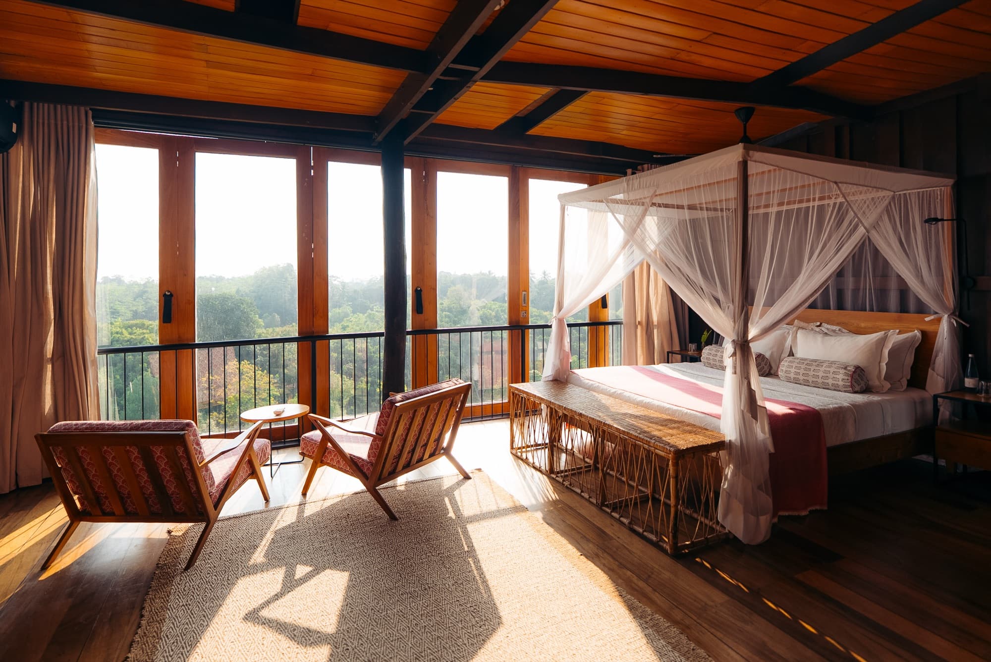 Bright king room with a canopy bed, two armchairs and floor-to-ceiling timber windows