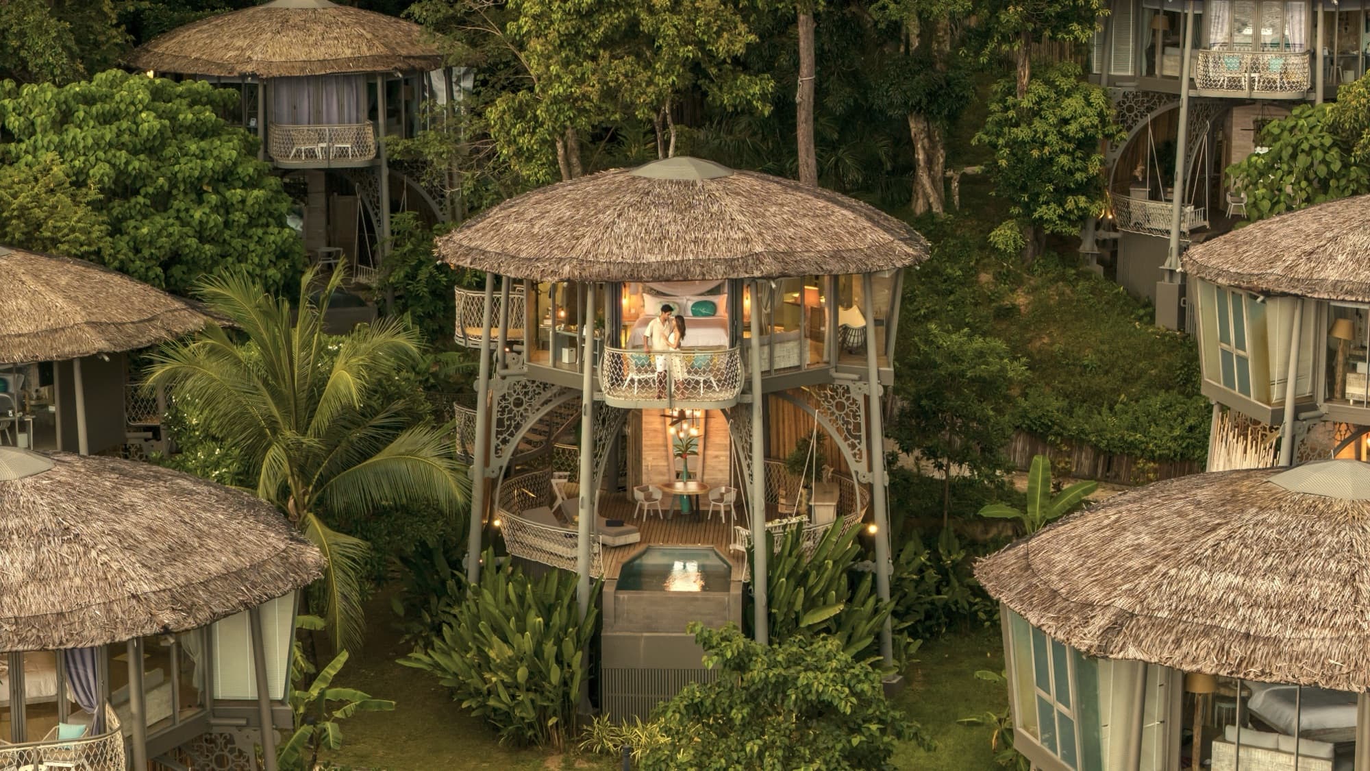 Aerial view at dusk of round thatched villas raised on stilts, lit from inside and surrounded by jungle