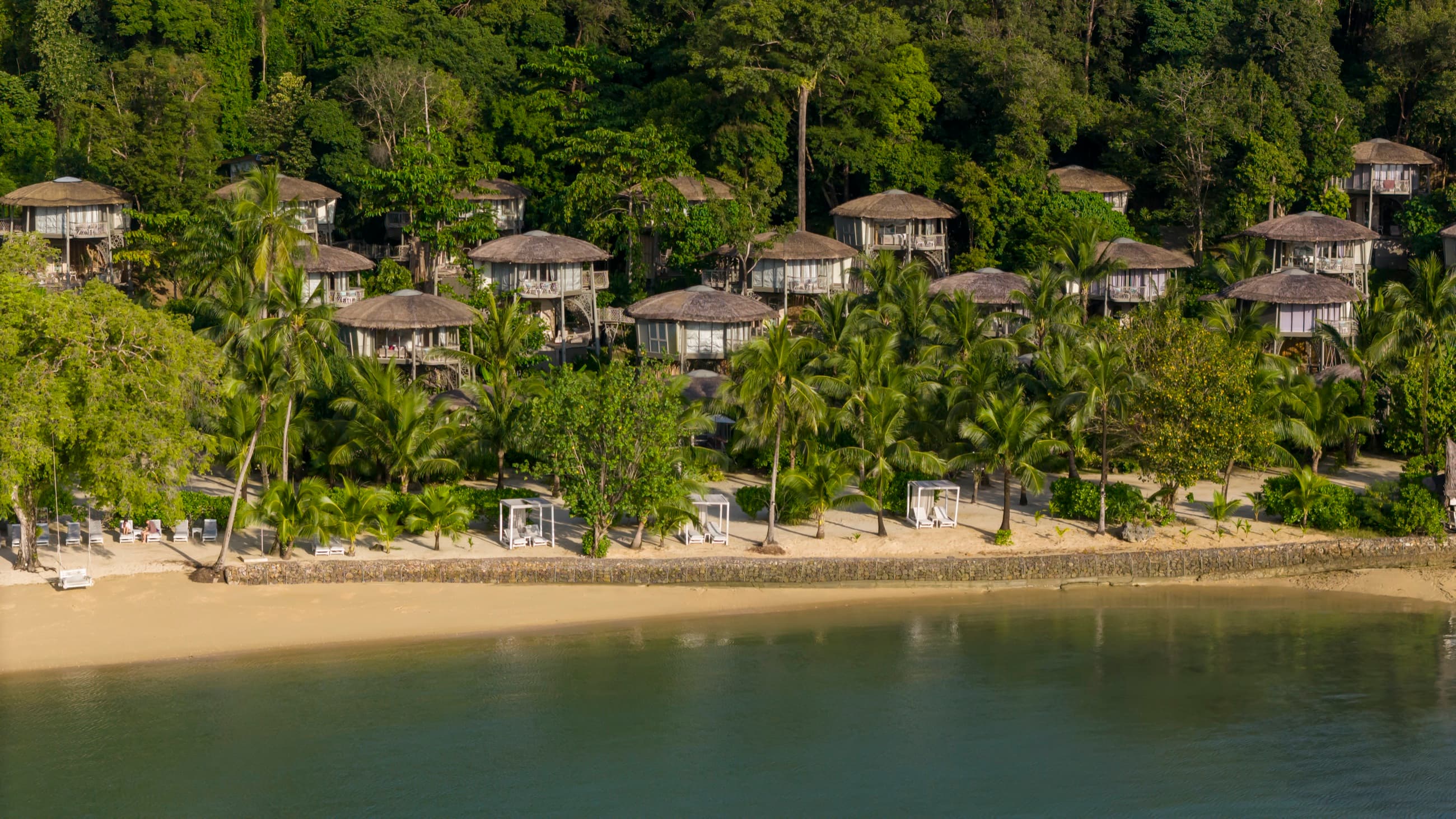 Thatched villas on stilts behind a curving sand beach, seen from the water, Koh Yao Noi, Phang Nga Bay