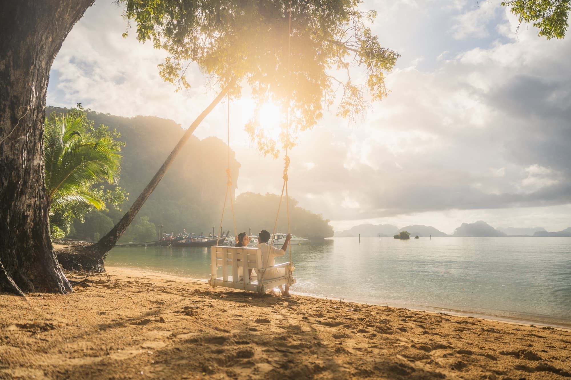 Wooden swing hanging from a leaning tree on the beach at sunrise, longtail boats moored offshore