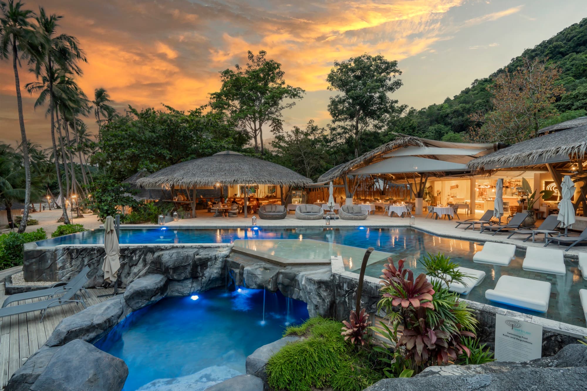 Clubhouse pool and the open-sided Roots restaurant lit at dusk, palms and forest behind