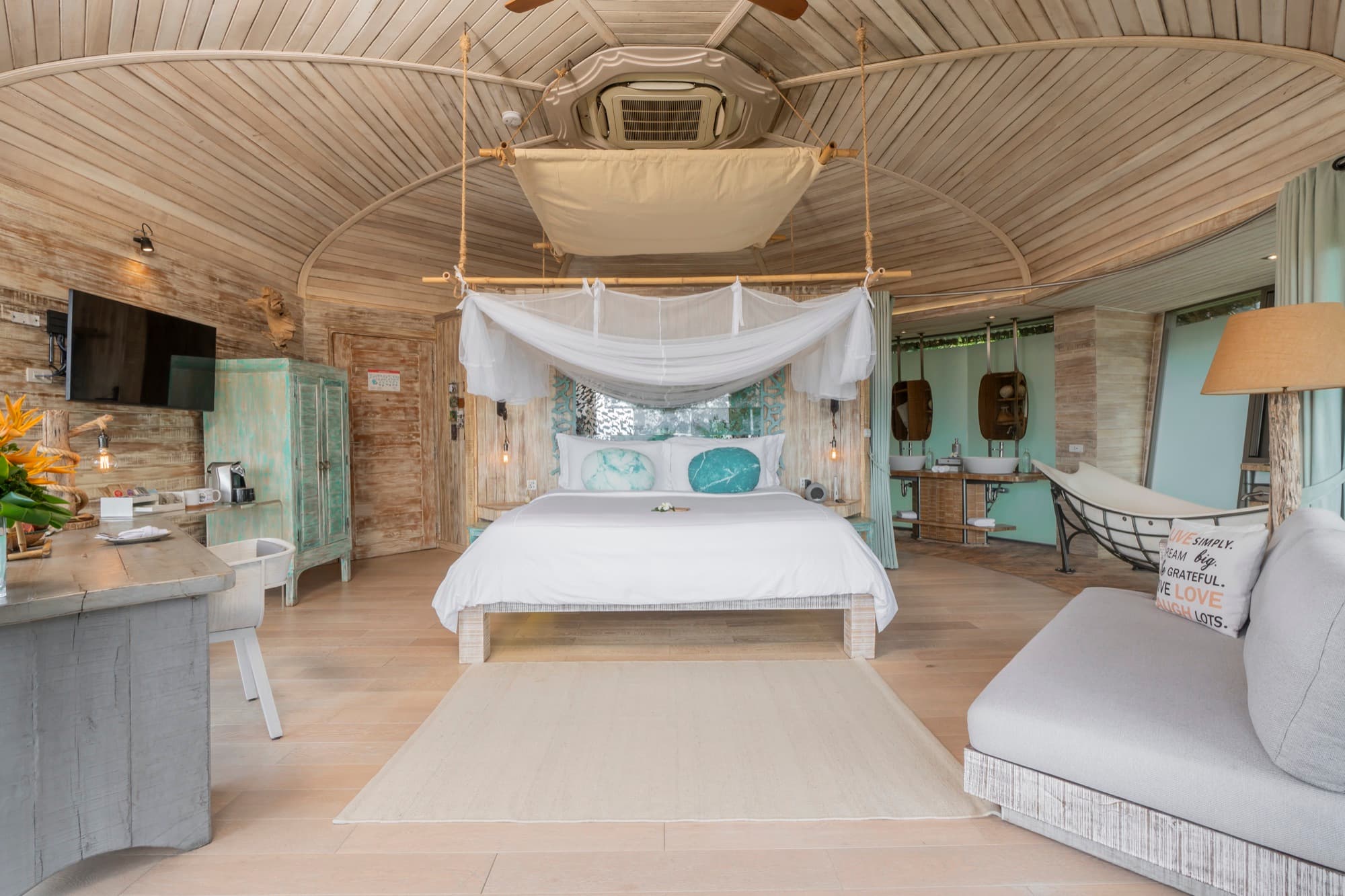 Villa interior with a canopy bed, a hanging boat-shaped daybed and an open bathroom under a domed wooden ceiling