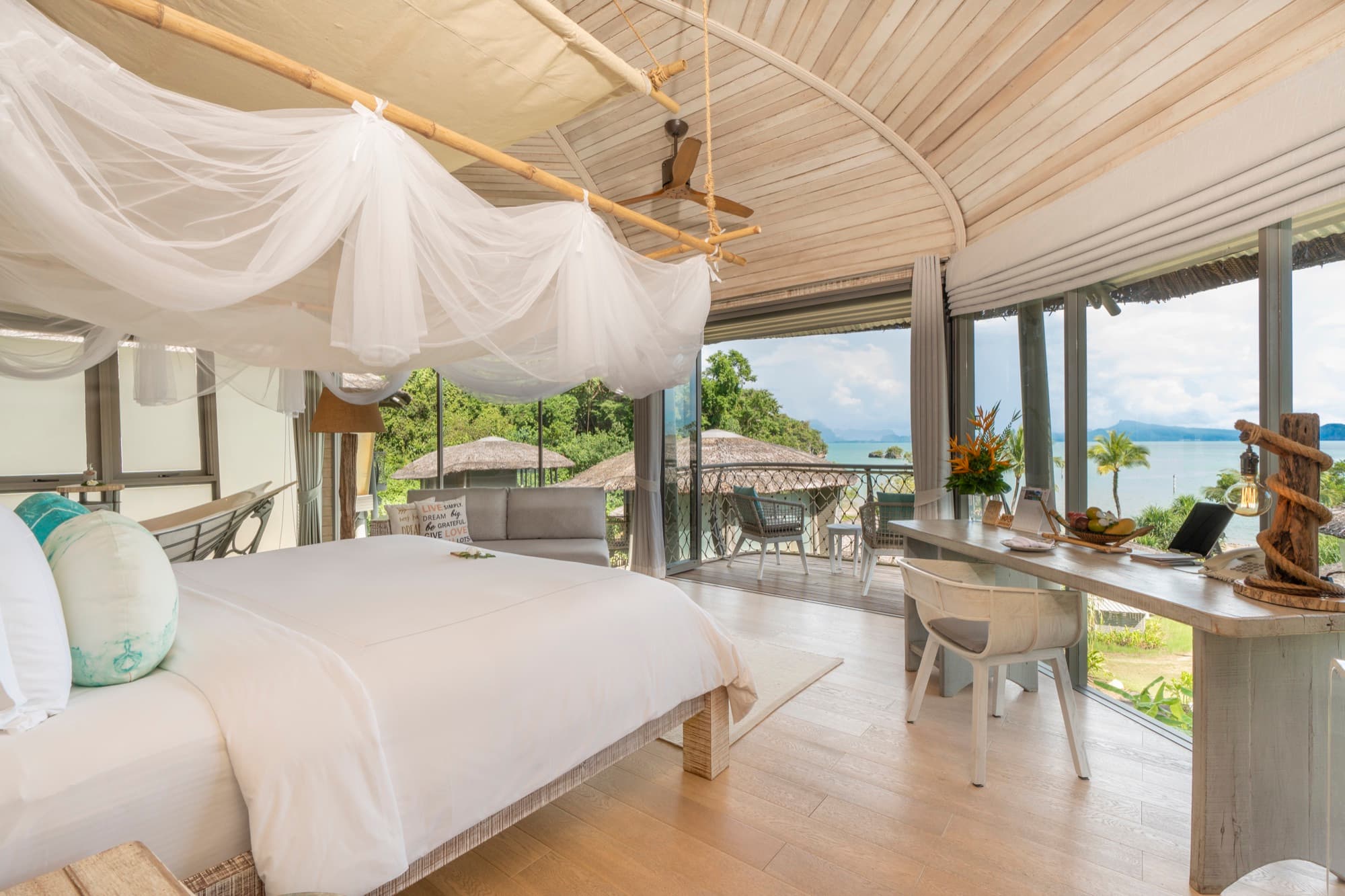 Bedroom under a curved timber ceiling, bed draped with a mosquito net, terrace and sea beyond the glass doors