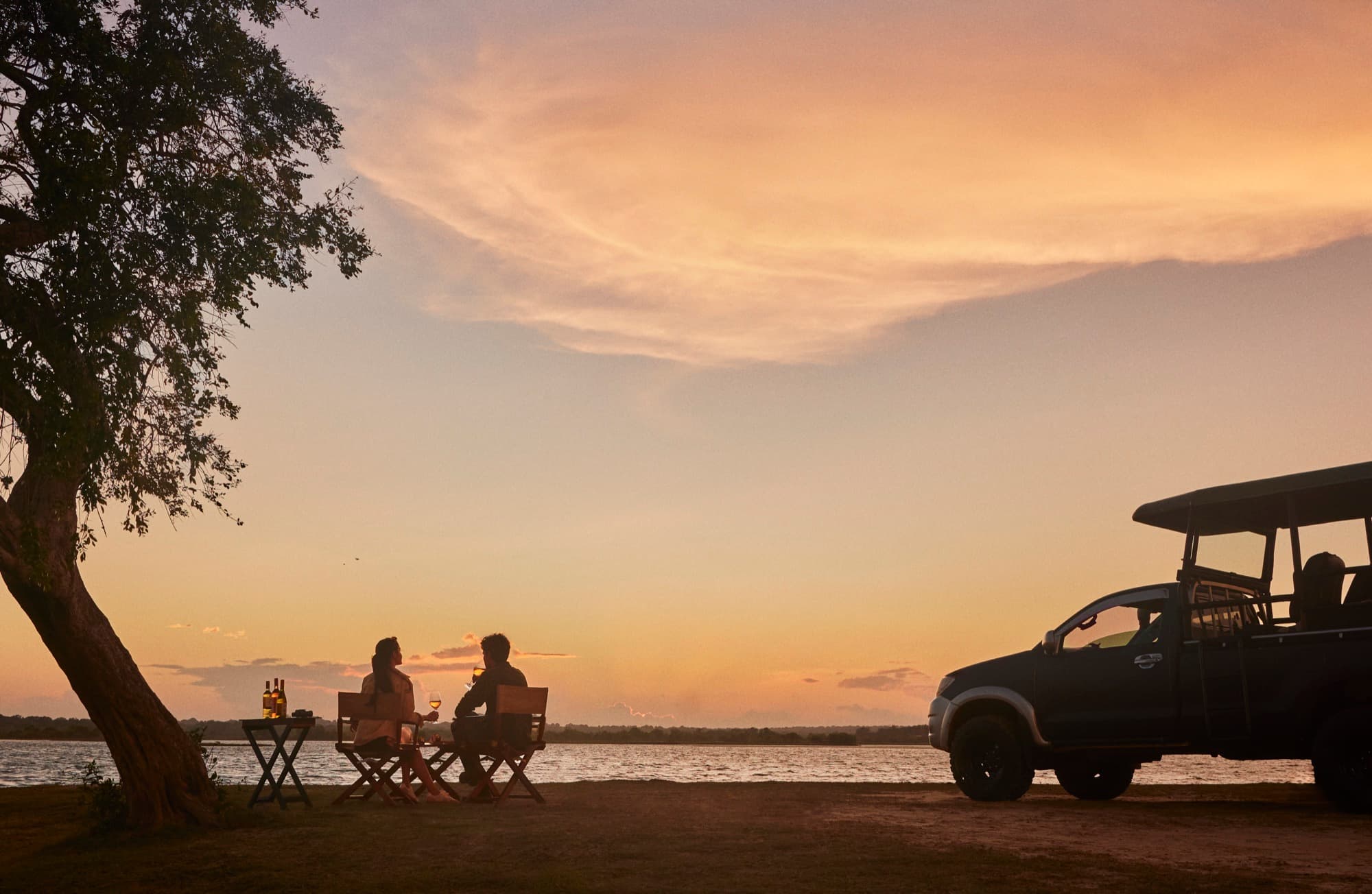 Guests at a table beside a safari jeep, by the water at sunset
