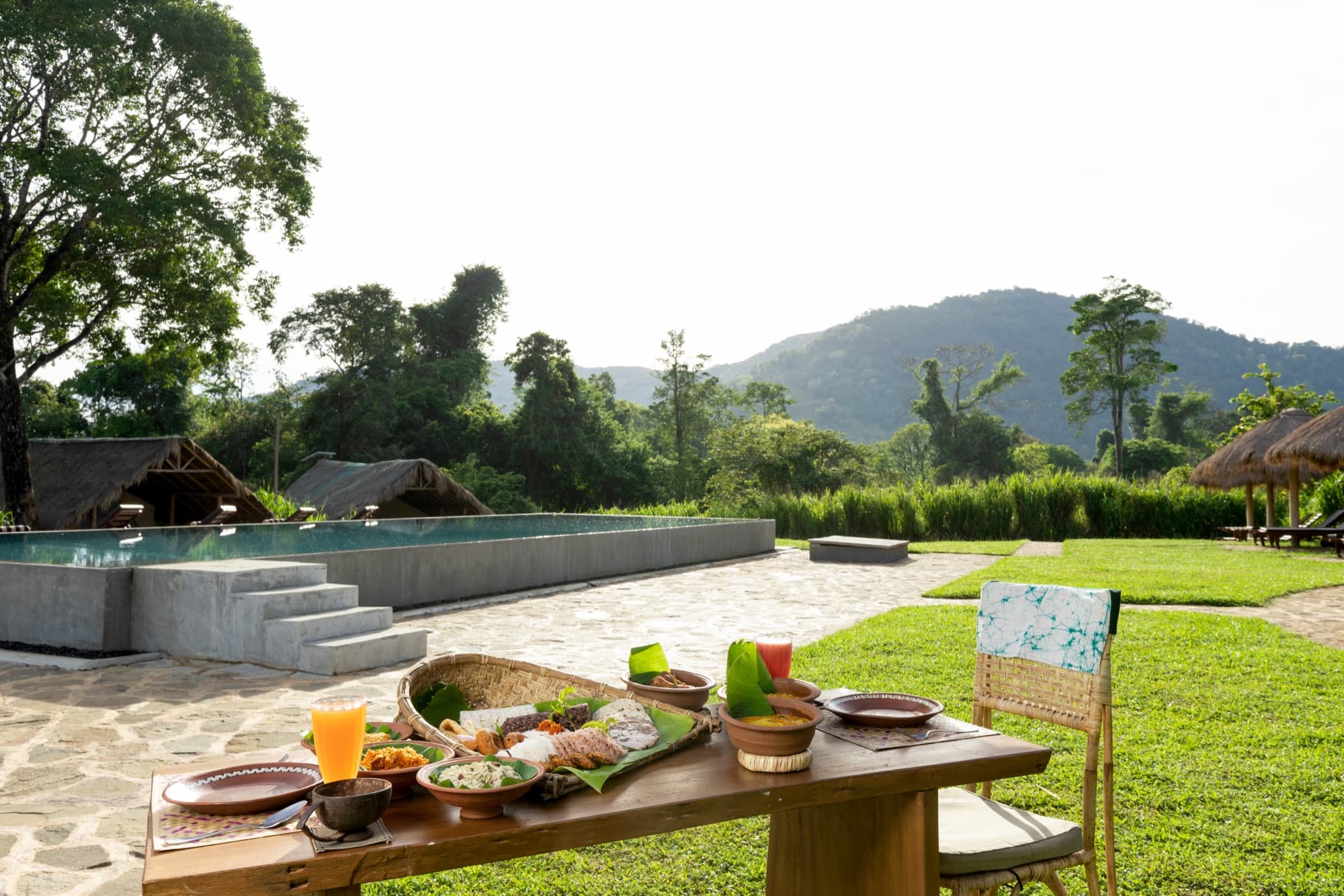 Open-air breakfast spread on a wooden table, pool and mountains in the background