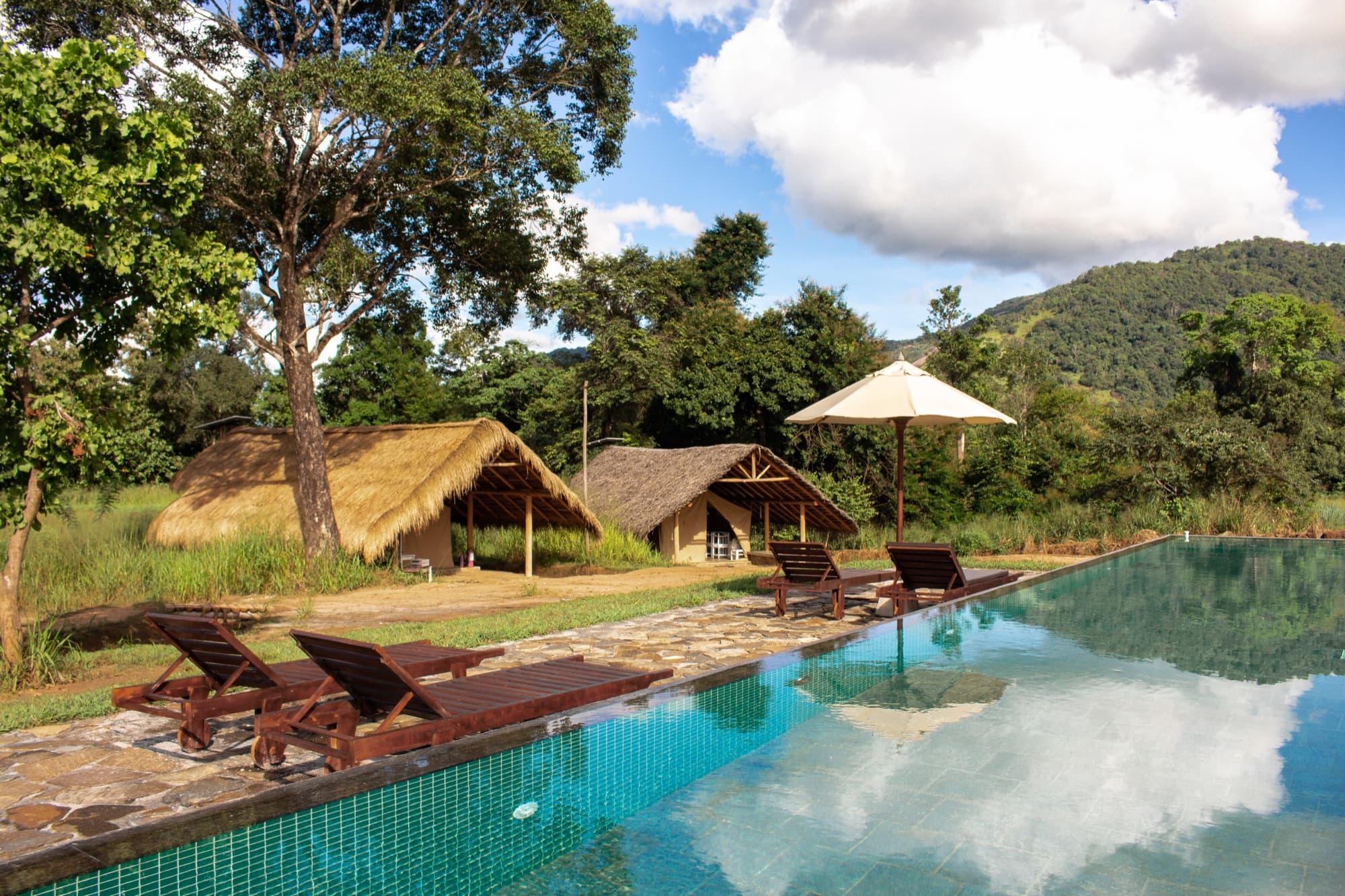 Infinity pool with sun loungers and umbrella facing the thatched tent pavilions and mountains