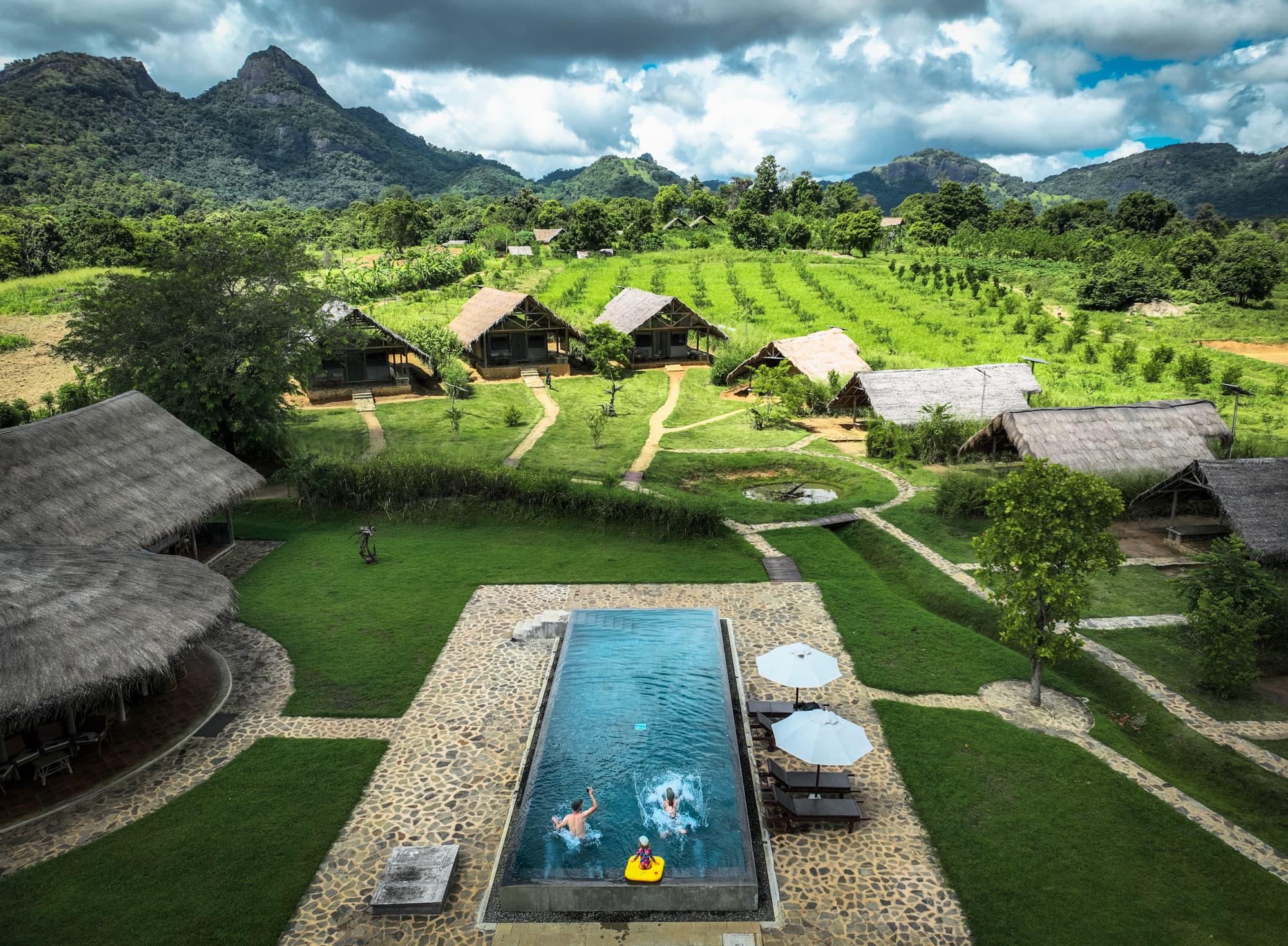 Aerial view of the infinity pool with swimmers, orchard rows and mountains behind the tented lodges