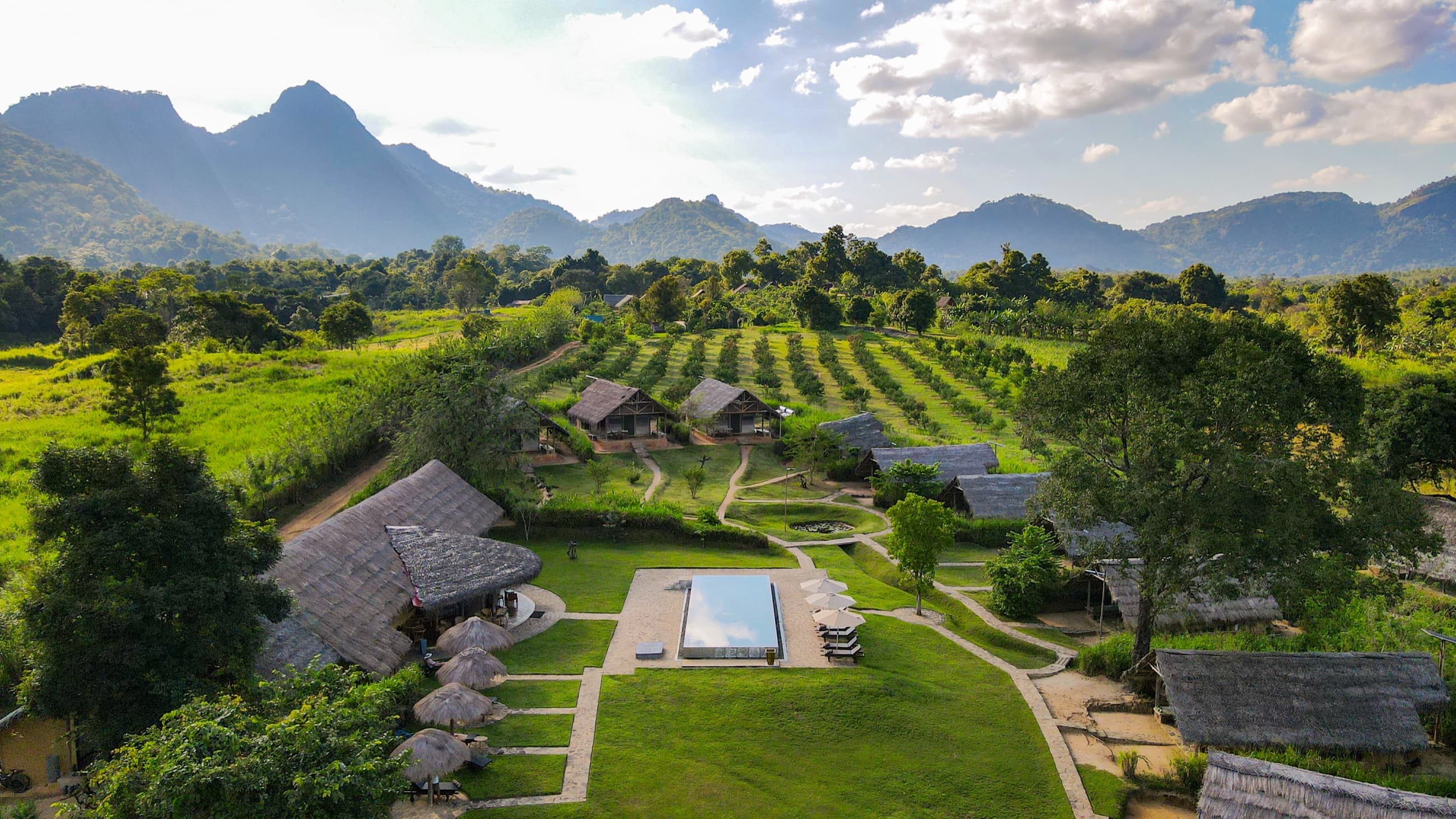 Aerial view of Wild Glamping Gal Oya — thatched pavilions, infinity pool and orchard rows below the mountains