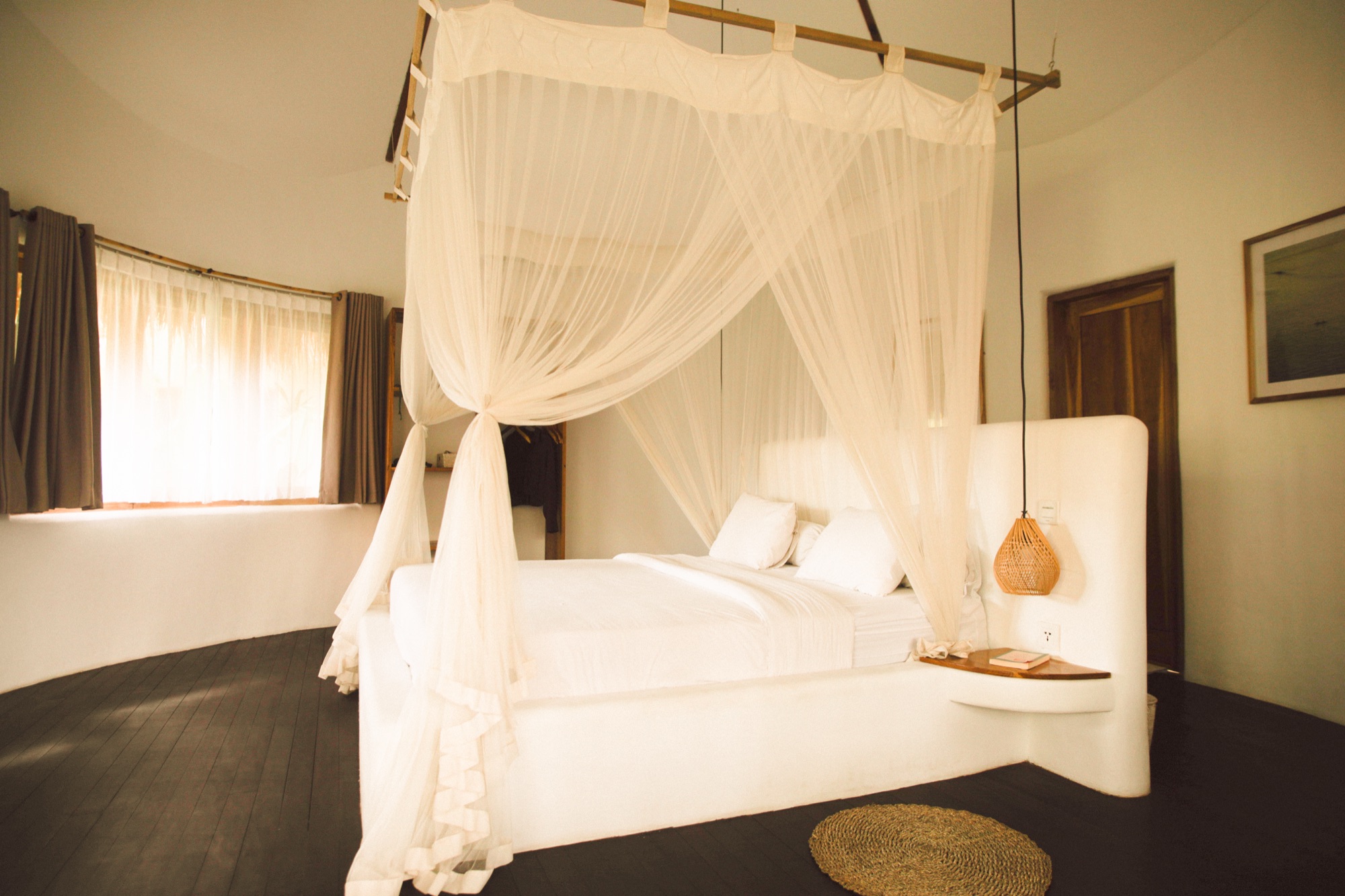 Room with a white canopy bed and mosquito net, dark timber floor and garden window