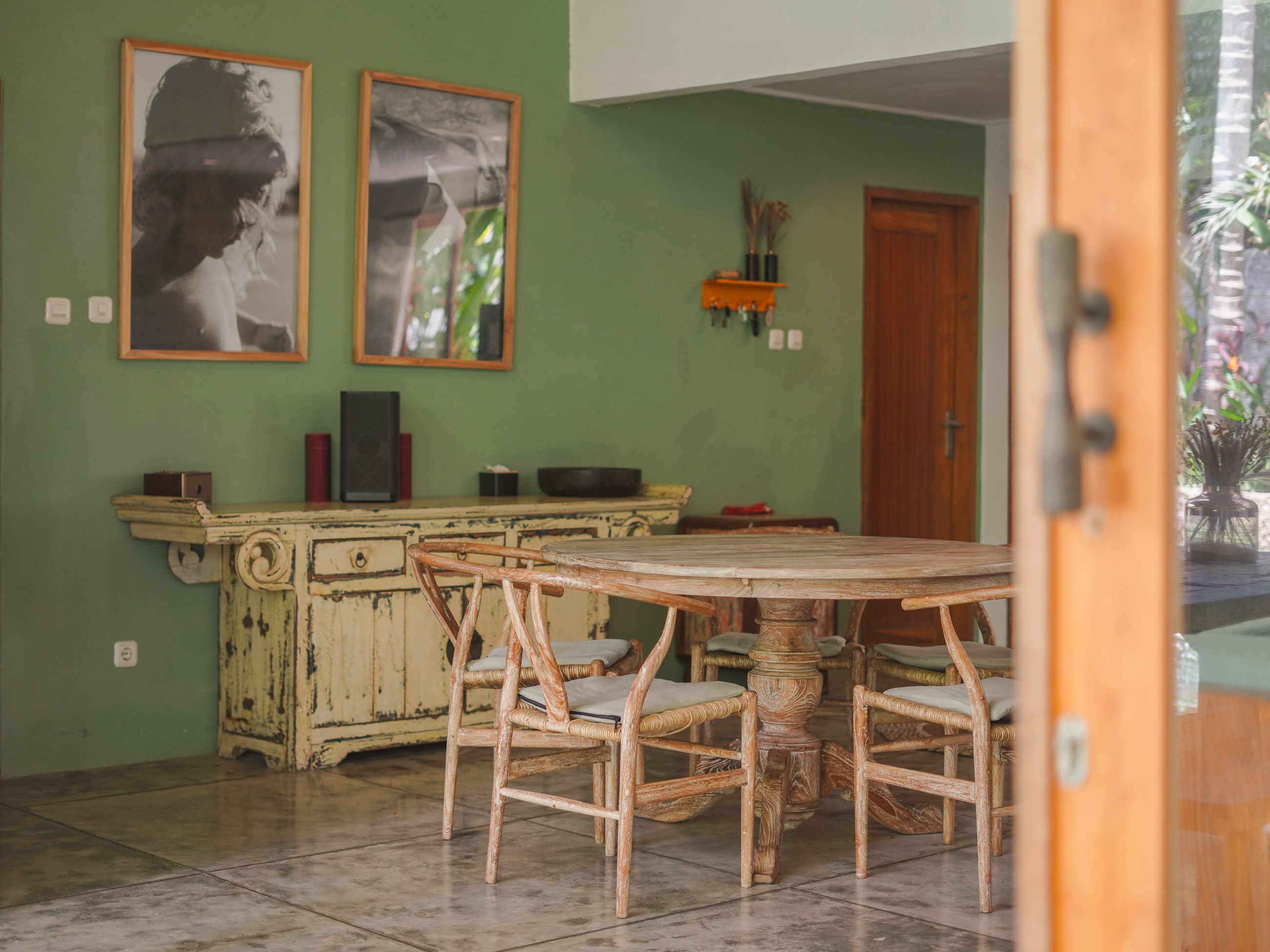 Villa dining corner with a round timber table, woven chairs and framed prints on a green wall