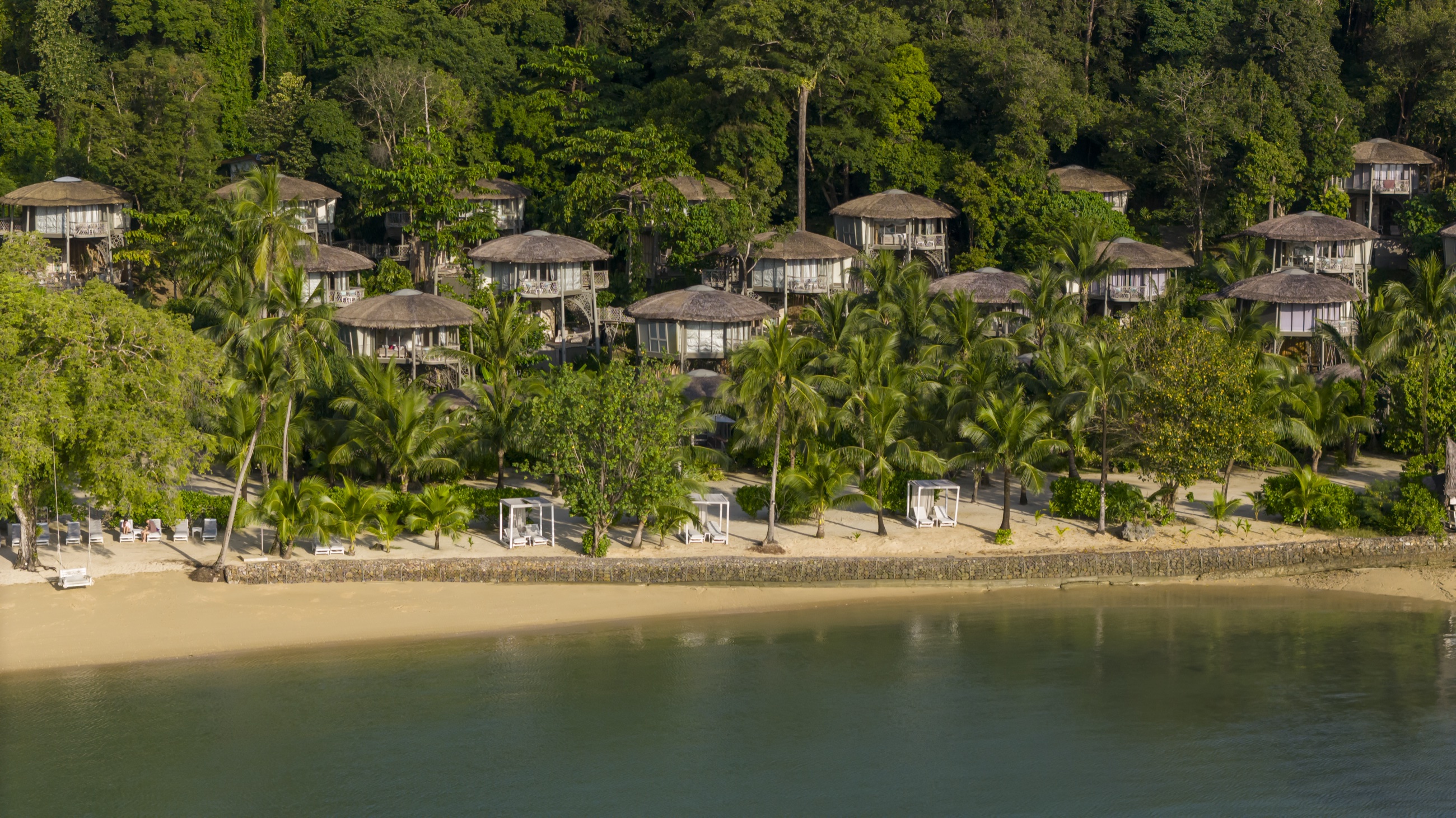 Thatched villas on stilts behind a curving sand beach, seen from the water, Koh Yao Noi, Phang Nga Bay