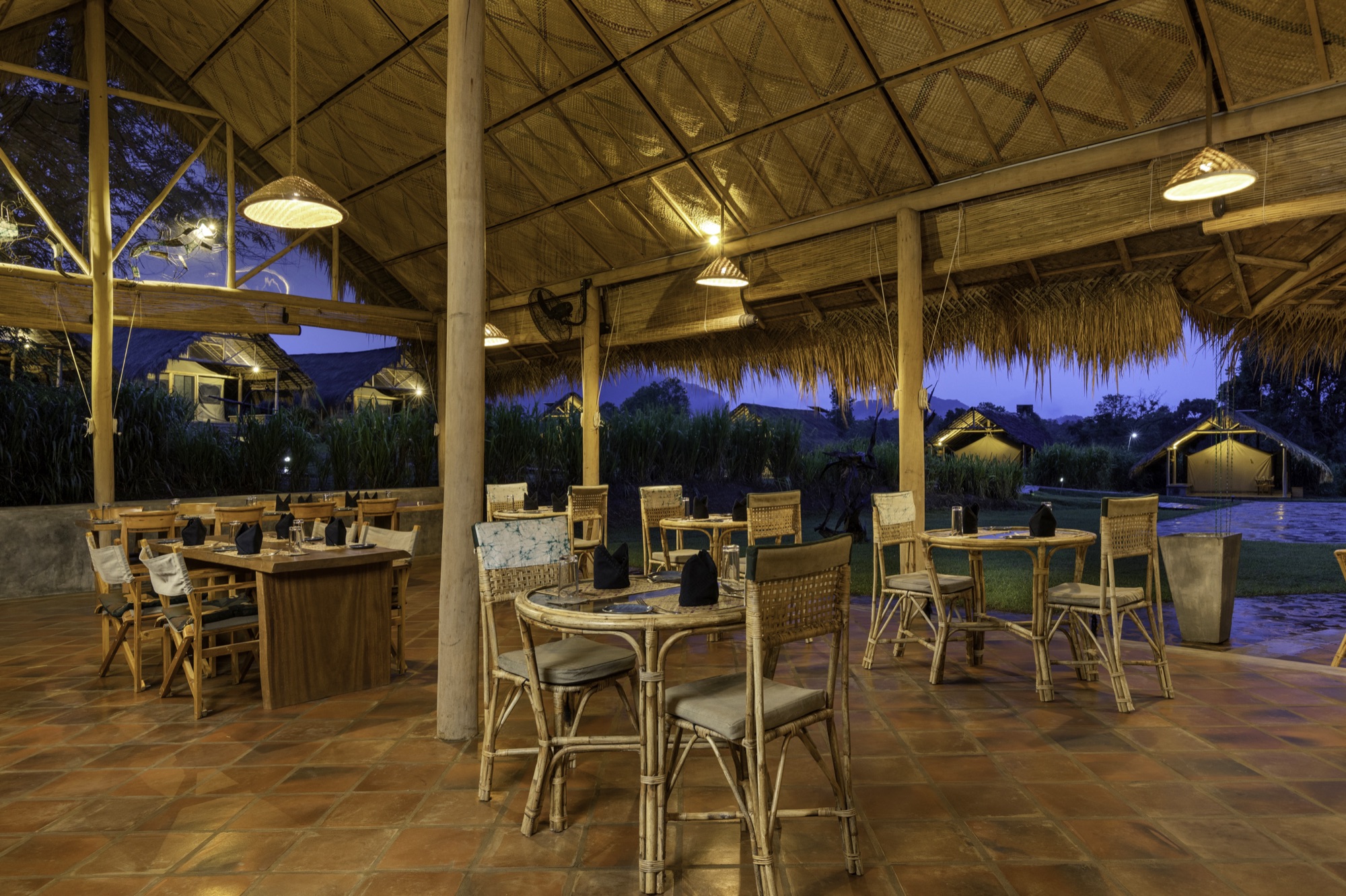 Main restaurant interior at dusk, lit lanterns under the thatched roof, tents visible outside
