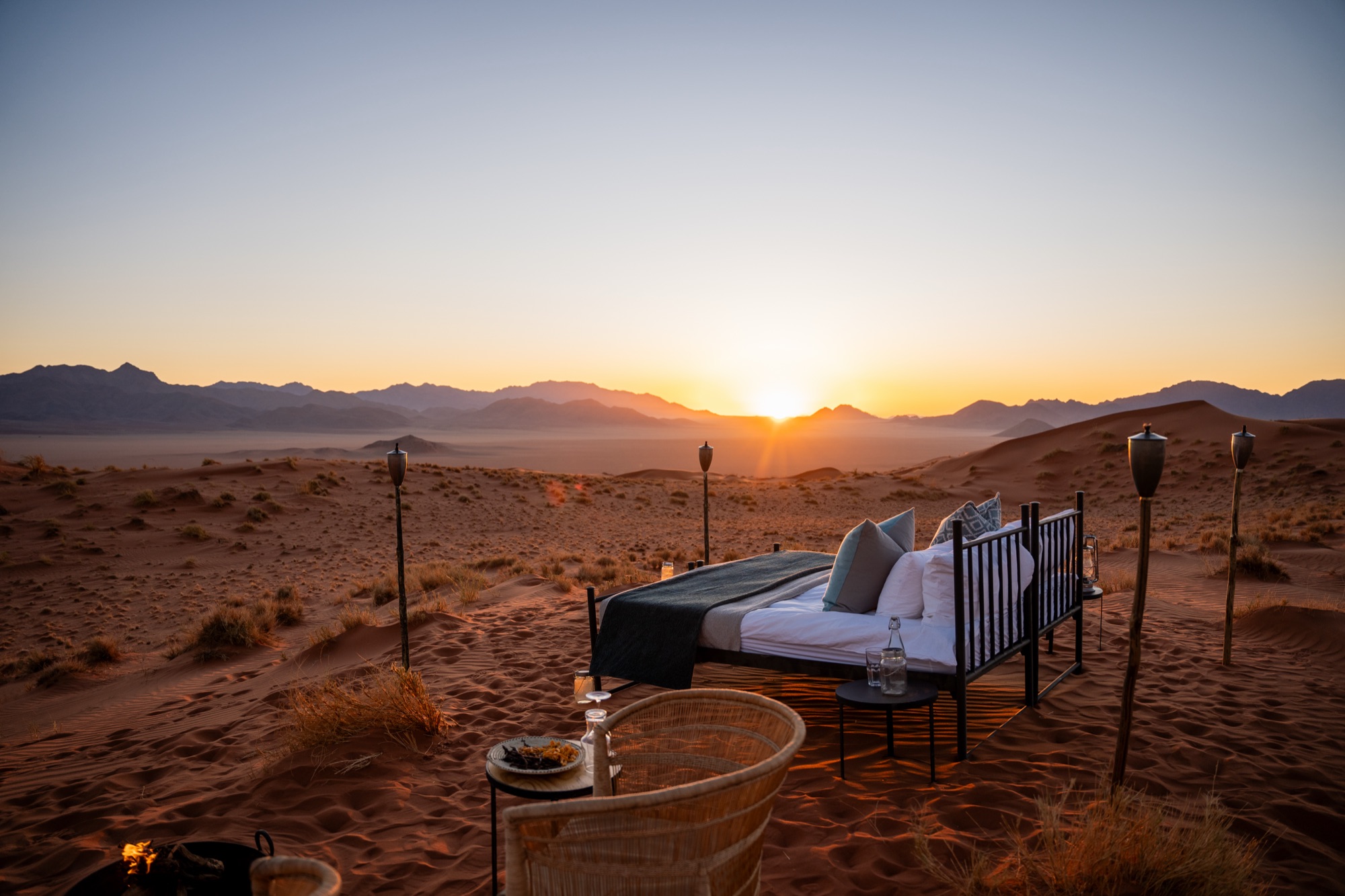 Bed set out in the desert at sunrise, between torches