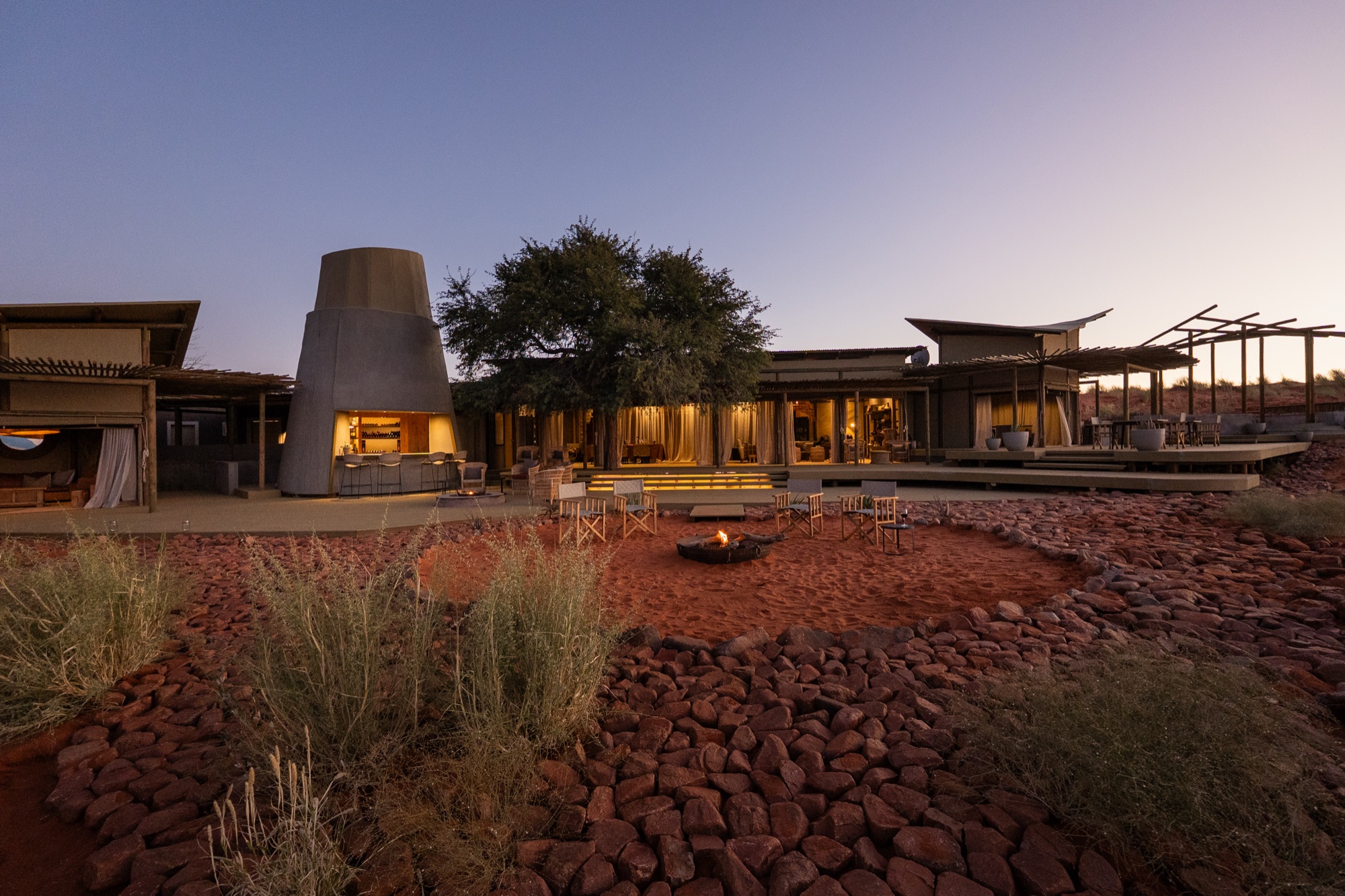 Main area at dusk, with the lit silo, a tree and the fire pit