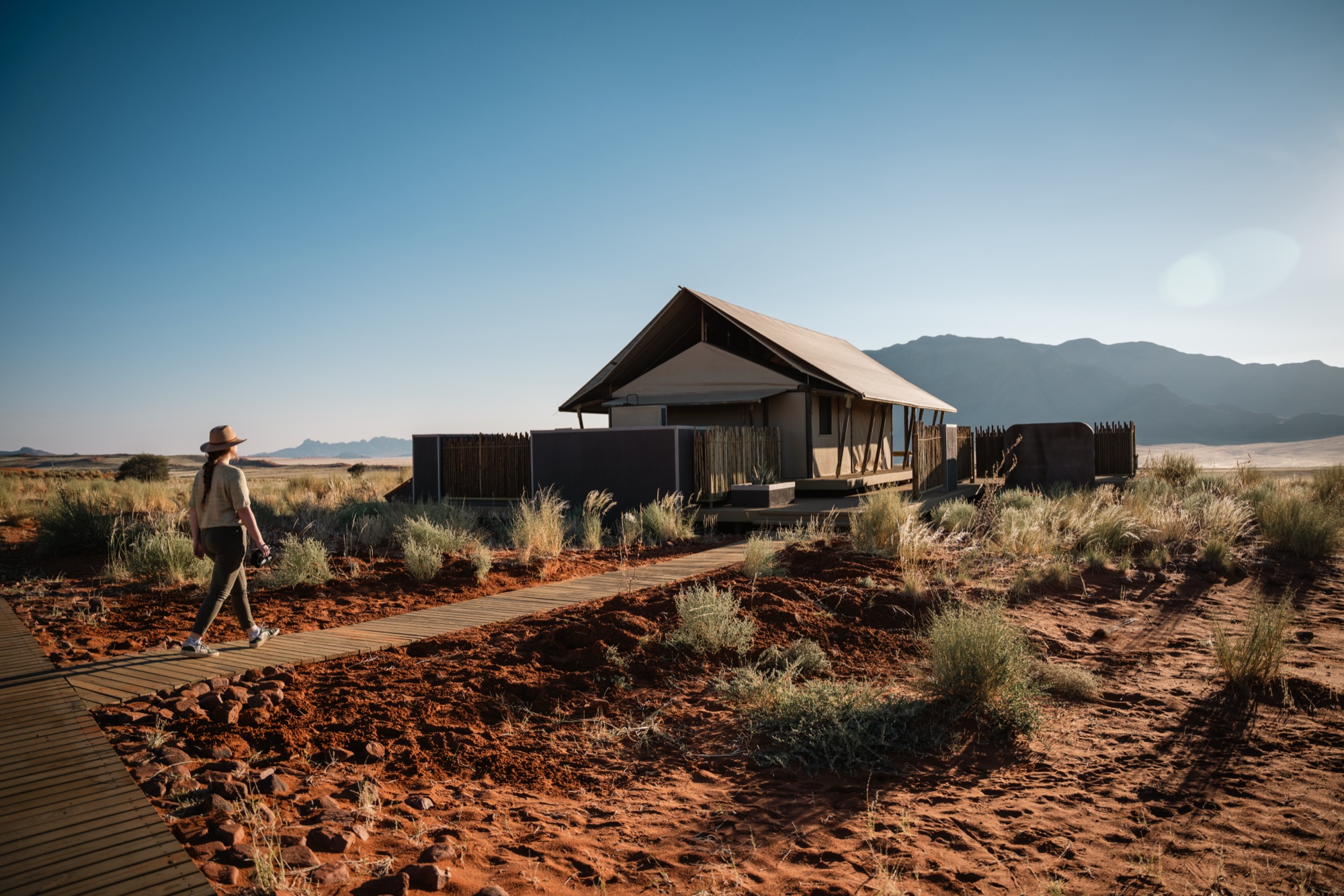 A guest walks the wooden boardwalk to a tented room, mountains behind